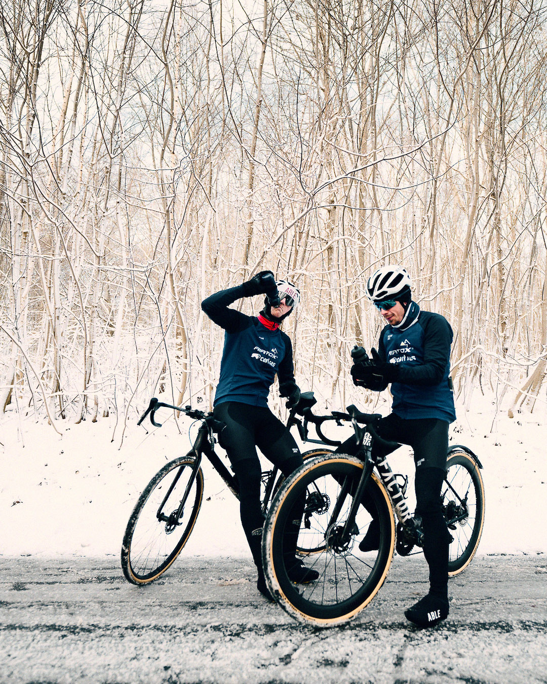 Two cyclists for Team Airtox standing together with their bikes on the snowy road.