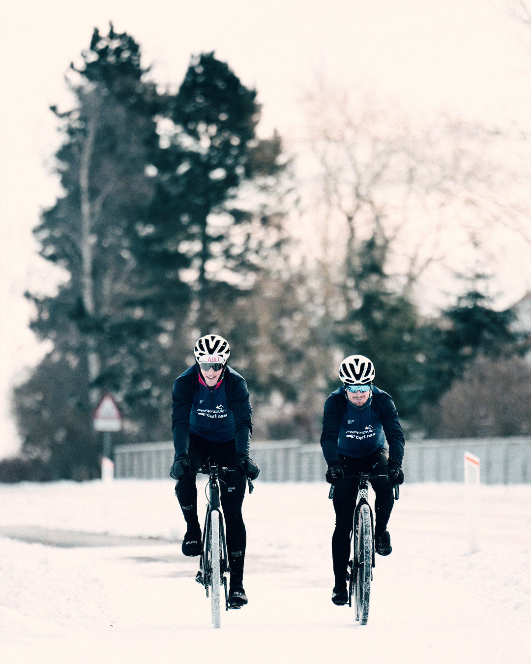 2 cyclists from Team Airtox cycling through the snowy road towards the camera. They are wearing Willing Able apparel.