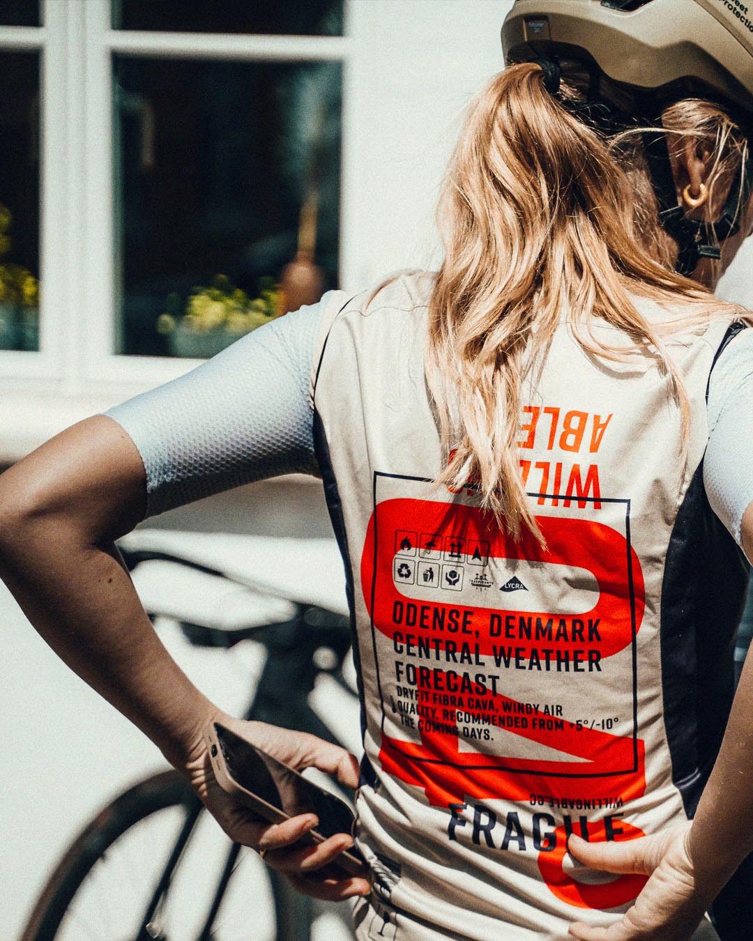 Woman cyclist from the back, wraing a WIlling Able shirt in a light white color and red color, with a helmet on.