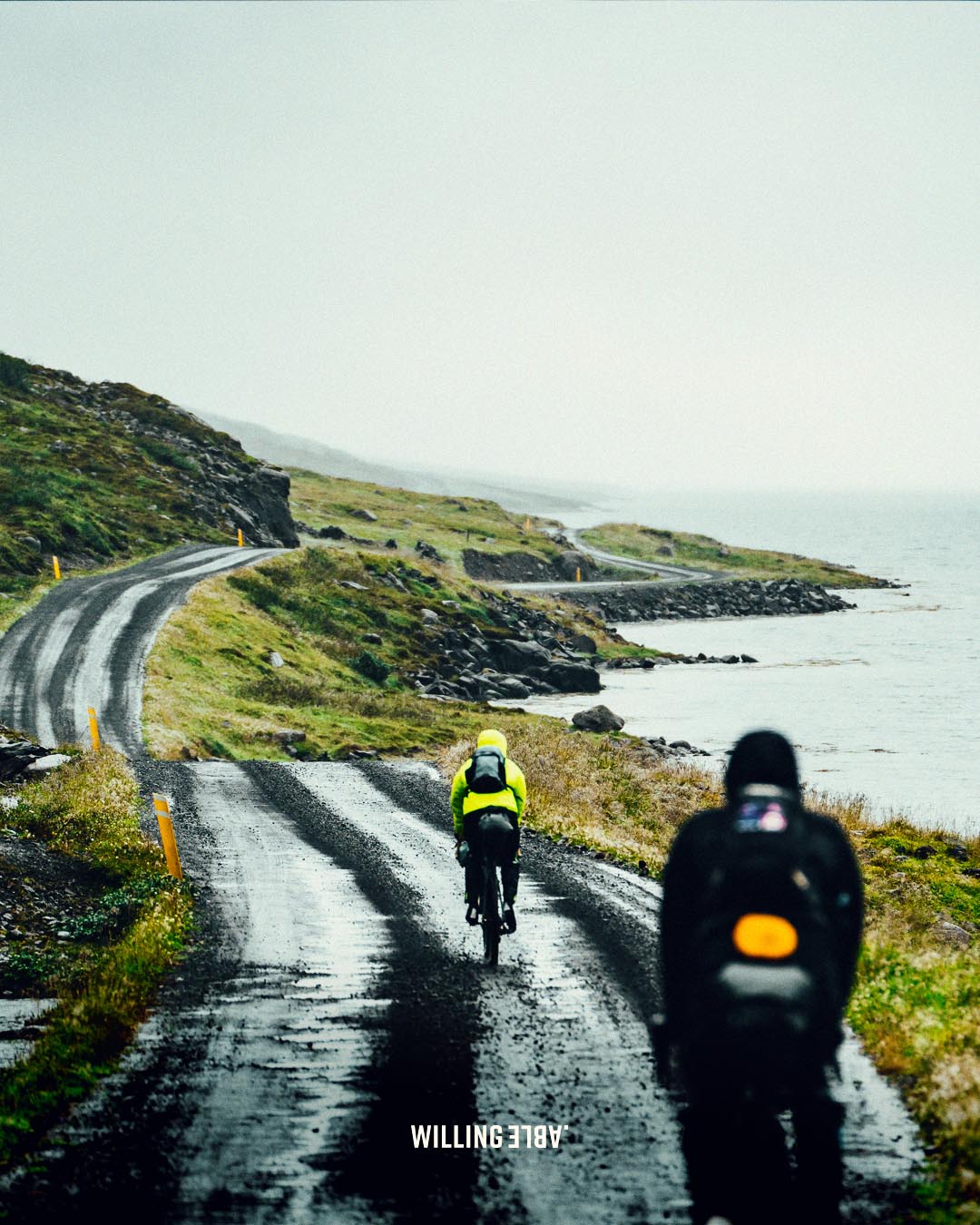 The two bikepackers cycling along a wet road through the nature og iceland, the sea to their right.