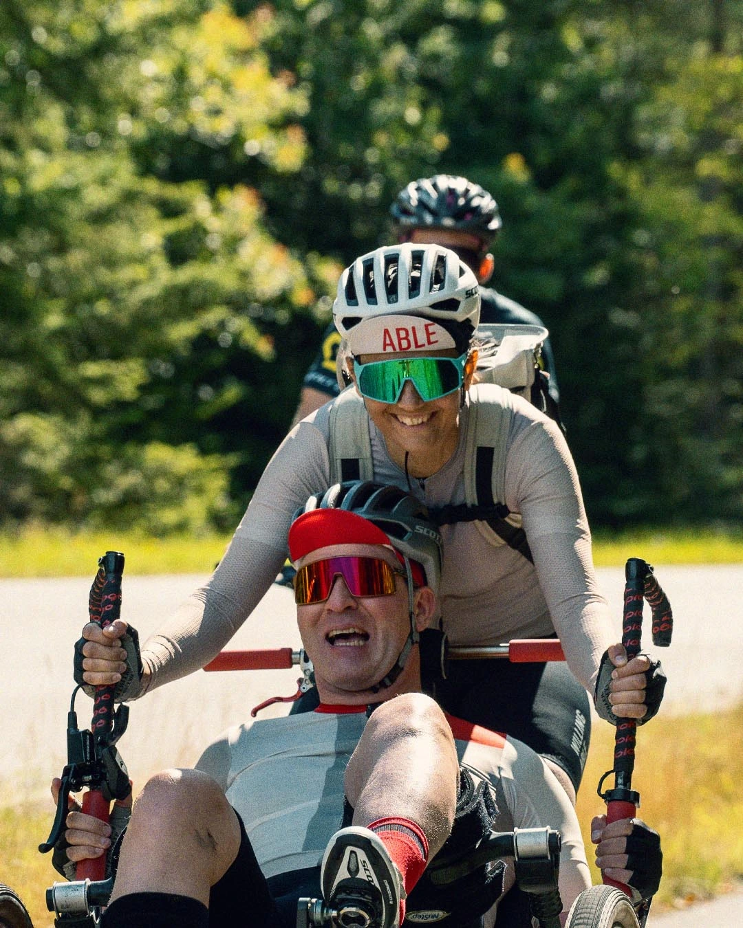 Michael Dvinge cycling on his three-wheeled bike with a female cyclist behind him. Both are smiling, and the woman is wearing a Willing Able hat. Both are cycling for people with disabilities.