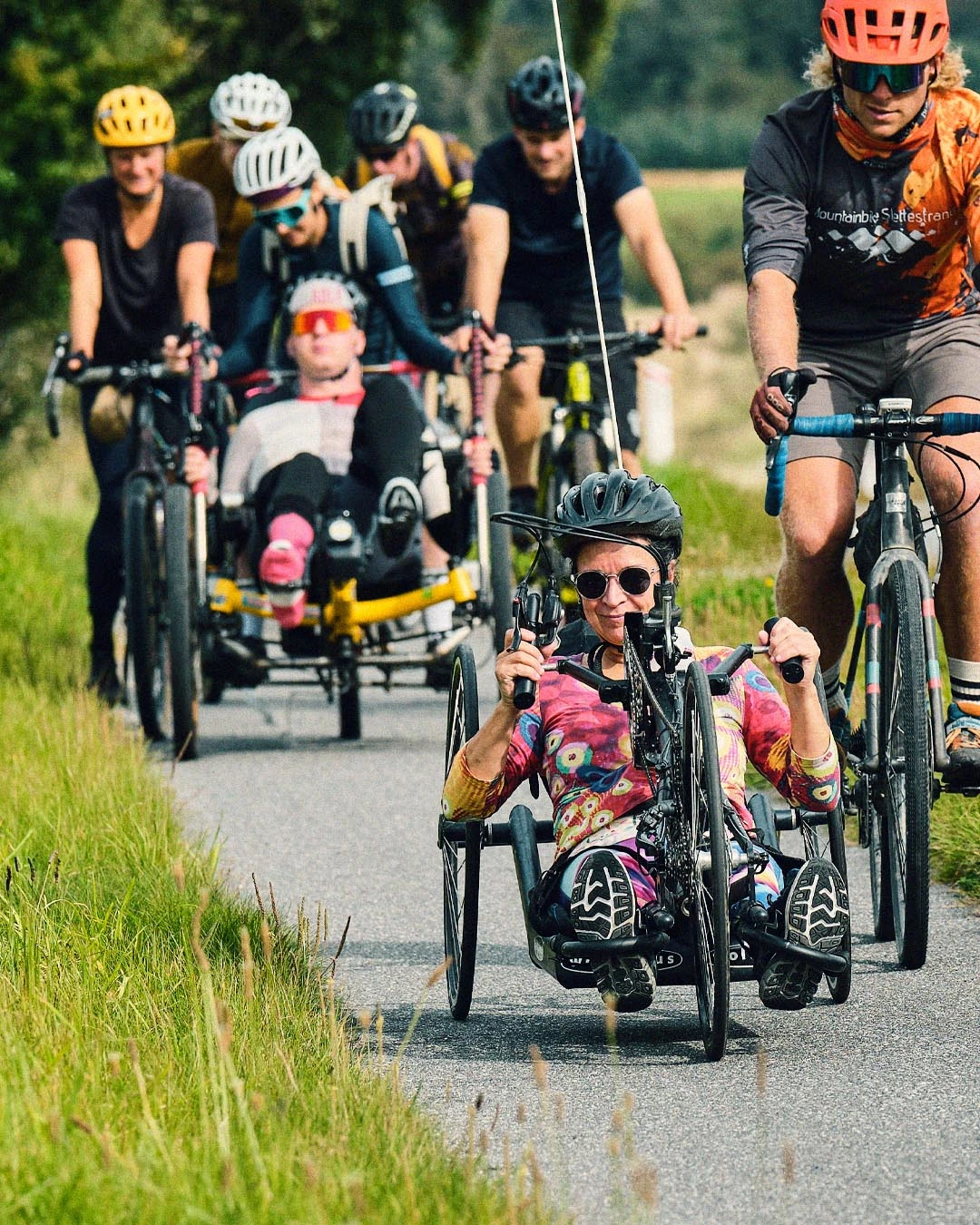 A woman with disabilities at the front on her three-wheeled bike in Tour de Jutland, with Michael Dvinge behind her on his three-wheeled recumbent bike, alongside other cyclists with and without disabilities on the road.