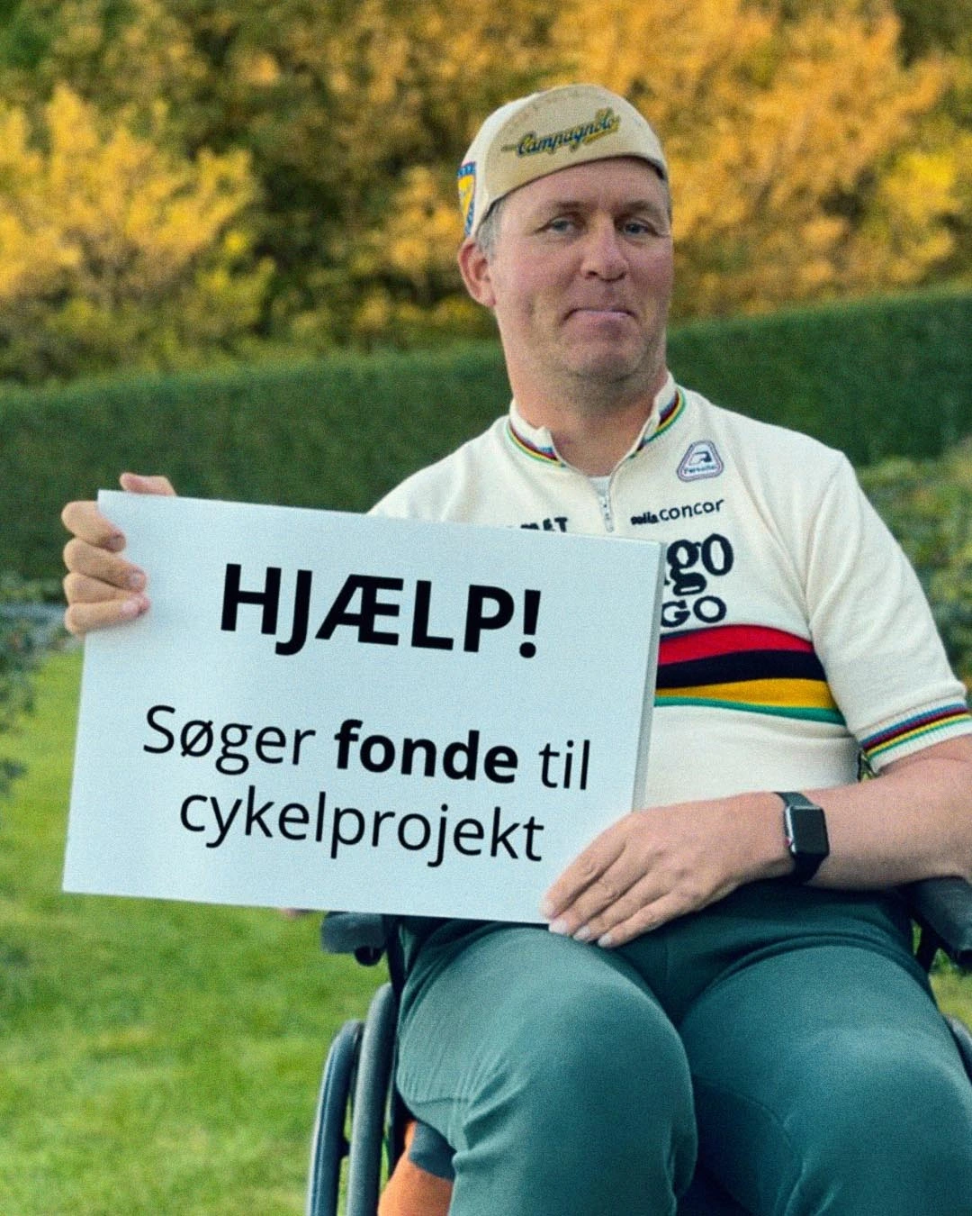 Michael Dvinge founder of Tour De Jutland smiling in his wheelchair outdoors, holding a sign that says, "Help! Seeking funding for cycling project.", inspiring others with disabilities.